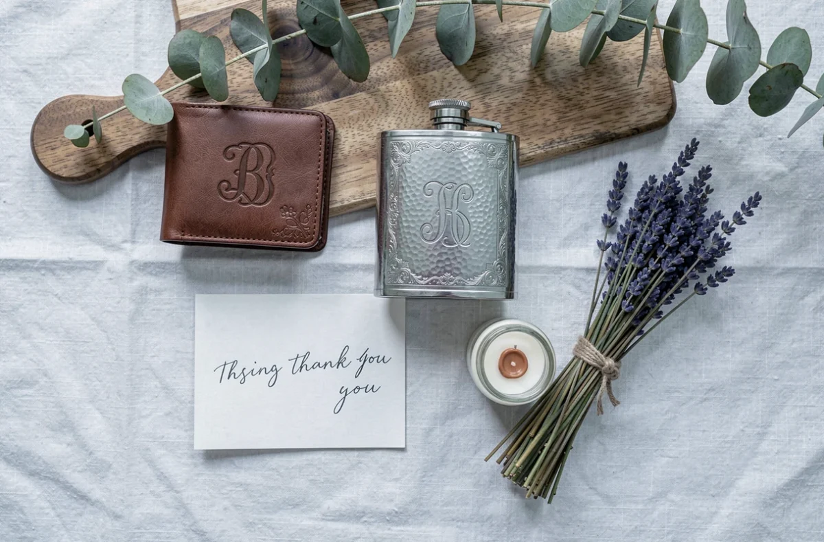 Flat lay de cadeaux intemporels pour témoins de mariage incluant portefeuille gravé, flasque argentée et carte manuscrite